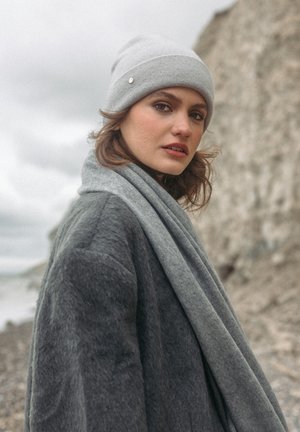 Grau Wollmütze mit silbernem Akzent, kombiniert mit einem grauen, strukturierten Schal und einem dunkelgrauen Kunstfellmantel, vor einer felsigen Küstenkulisse.