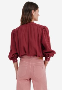 Blouse bordeaux avec des manches bouffantes, en tissu texturé, et des poignets à boutons, associée à un jean taille haute rose pâle et à un détail d'étiquette.