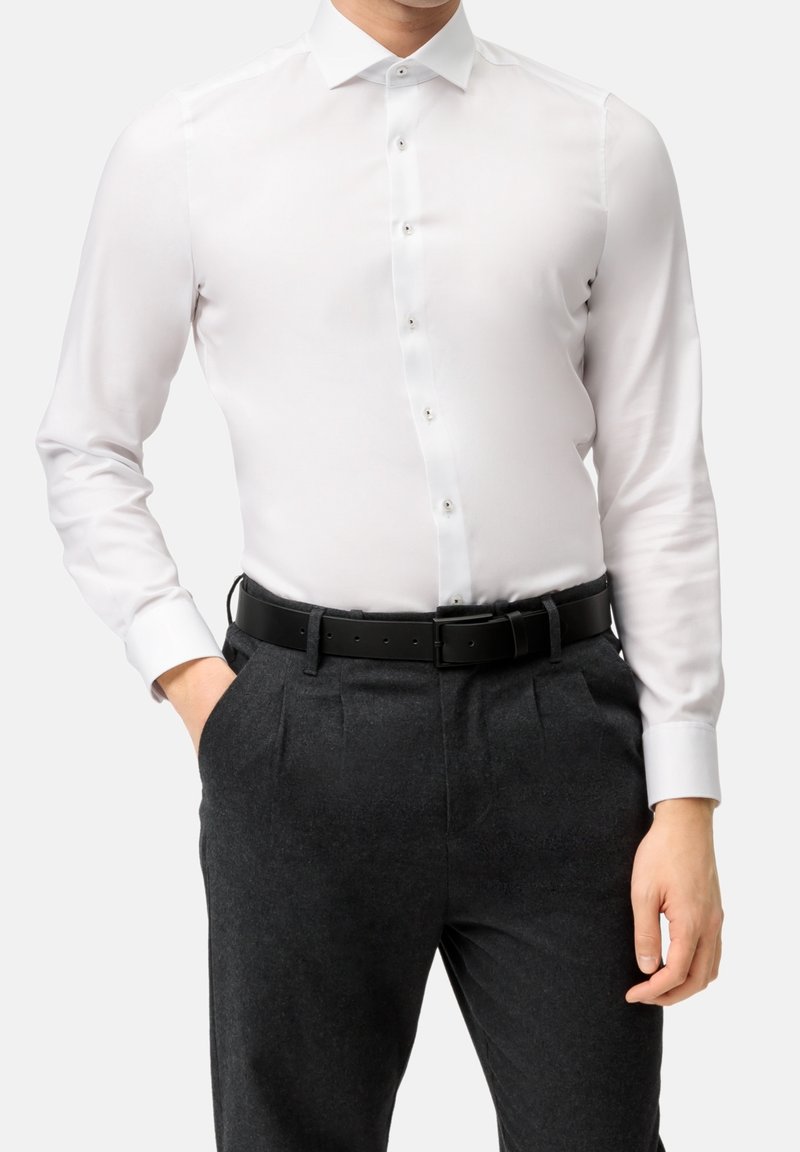 Homme portant une chemise blanche ajustée rentrée dans un pantalon plissé gris foncé avec ceinture noire, une main dans la poche, sur fond uni.