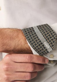 Kragenhemd aus strukturiertem weißen Stoff mit einem gemusterten dunklen Manschette, akzentuiert in Gold und Weiß, das ein geometrisches Design zeigt.