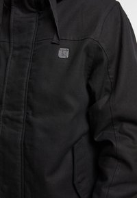 Chaqueta negra con un tejido texturizado, que presenta una capucha, un bolsillo frontal y un pequeño logo cuadrado en el pecho. Diseño simple y funcional.