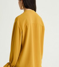 Blusa giallo senape con maniche lunghe, dettagli arricciati ai polsi, texture liscia e vestibilità comoda. Semplice scollatura rotonda con una cucitura sul retro.