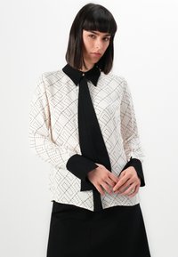 Long-sleeve blouse in cream with a black collar and cuffs. Features a black tie detail and a geometric diamond pattern in red.