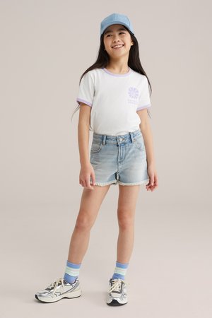 Uśmiechnięta dziewczyna w niebieskiej czapce, białej koszulce z fioletowymi wykończeniami, dżinsowych szortach, paskowanych skarpetkach i srebrnych trampkach stoi na jednolitym tle.