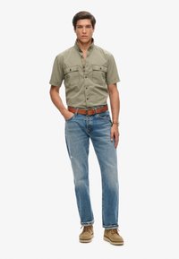 Camisa verde oliva de manga corta con bolsillos abotonados, combinada con vaqueros azul claro y zapatos beige. Un cinturón de cuero marrón añade contraste.