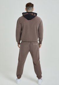 Conjunto de sudadera y pantalones de chándal marrón con capucha negra, que presenta el texto "SIRKIK". Fabricado con una tela suave, con puños acanalados y bolsillos laterales.