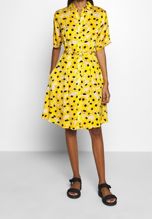 Mujer con vestido tipo camisa hasta la rodilla de flores amarillas, de manga corta, con cinturón en la cintura y sandalias negras, de pie frente a un fondo liso.