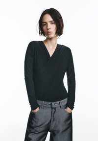 Čierne body s dlhým rukávom a zavinovacím dizajnom, vyrobené z mäkkej, pružnej látky. Kombinované so sivými, voľne strihanými nohavicami so zapínaním na gombíky.