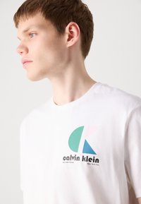 Balta medvilninė marškinėliai su daugiaspalviu logotipu, kuriame dominuoja žalia, rožinė ir mėlyna. Apvalus kaklas, trumpomis rankovėmis ir laisvas tinkamas.