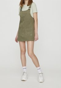 Robe salopette en jean kaki avec deux poches avant et bretelles ajustables, portée sur un t-shirt rayé, associée à des baskets blanches épaisses.