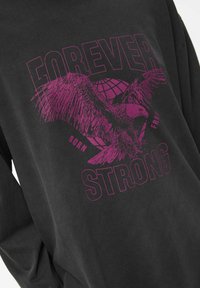 Czarna koszula z długim rękawem z dużym fioletowym wzorem orła oraz napisem "FOREVER STRONG" i wzorem globu. Miękka struktura tkaniny.