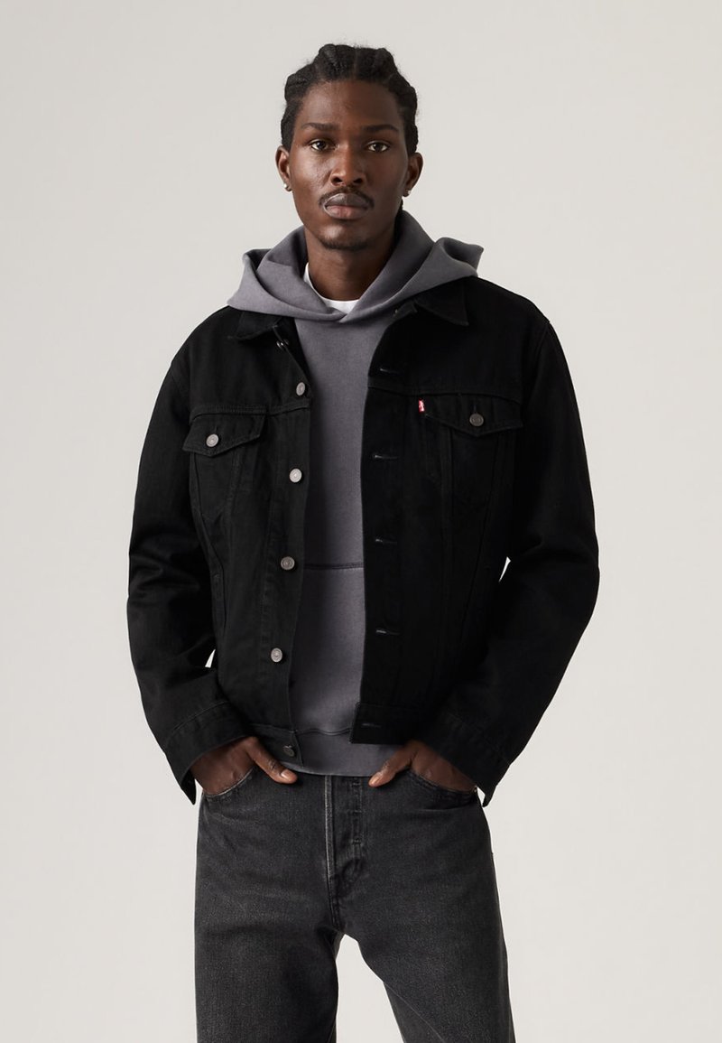 Black denim jacket with classic design, metal buttons, and two chest pockets, layered over a gray hoodie, paired with dark jeans.