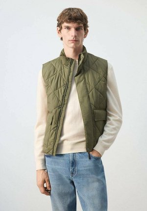 Joven con chaleco acolchado verde oliva sobre suéter crema y jeans azules, de pie con una mano en el bolsillo contra un fondo liso.