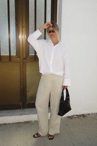 Weißes, übergroßes Hemd mit Knöpfen, beigefarbene, weite Leinenhosen, schwarze Sandalen und eine kleine schwarze Handtasche, stehend neben einem braunen Fensterrahmen.