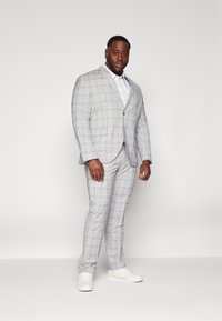 Traje a rayas gris con un corte ajustado, que cuenta con una chaqueta de un solo botón y pantalones a juego. Combinado con una camisa blanca y zapatillas blancas.