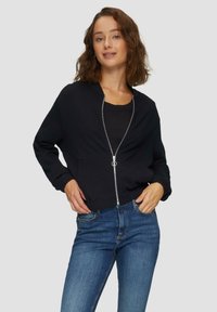 Schwarze Zip-Jacke mit gerippter Oberflächenstruktur, rundem Halsausschnitt und elastischen Bündchen, kombiniert mit dunkelblauen Jeans in einer lässigen Pose vor einem grauen Hintergrund.