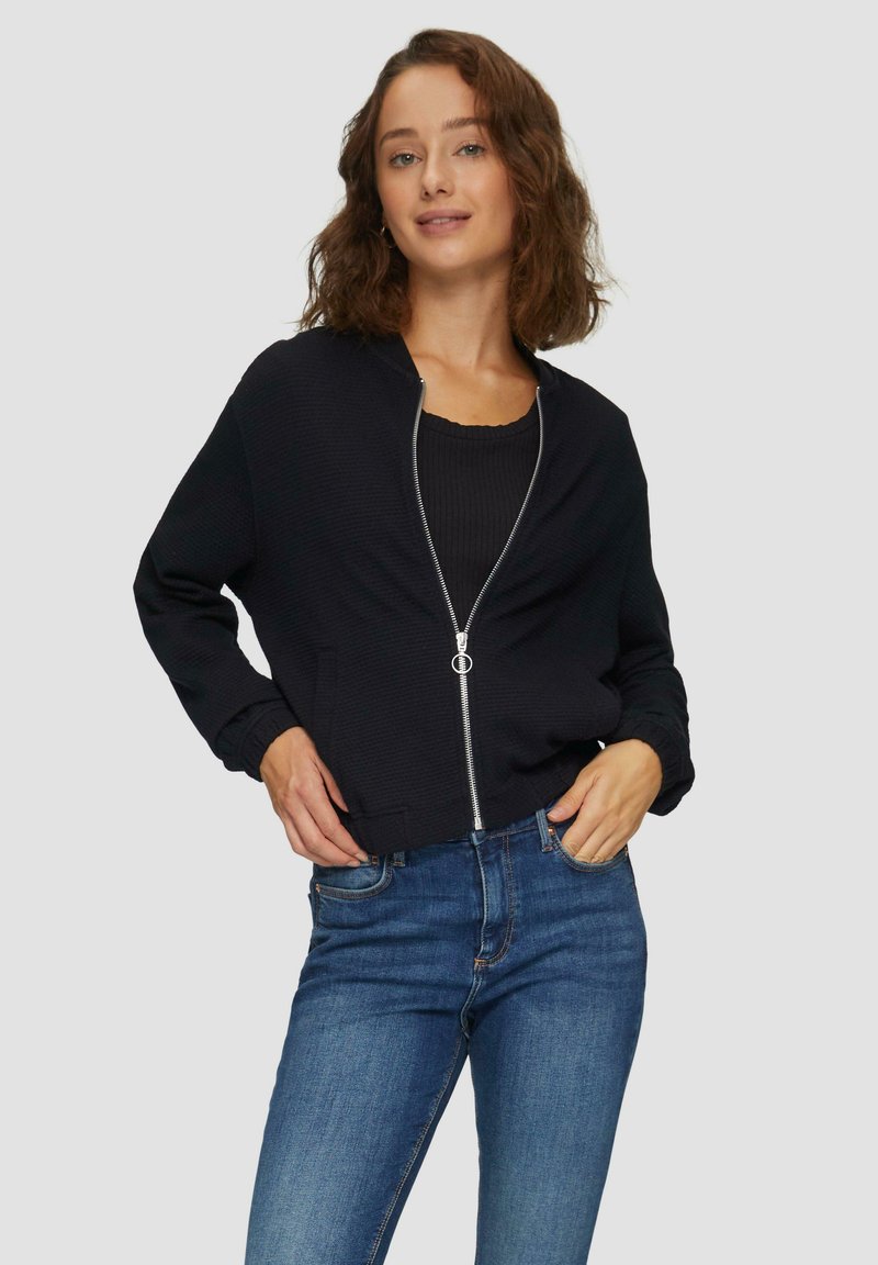 Schwarze Zip-Jacke mit gerippter Oberflächenstruktur, rundem Halsausschnitt und elastischen Bündchen, kombiniert mit dunkelblauen Jeans in einer lässigen Pose vor einem grauen Hintergrund.