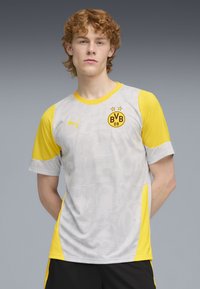 Jersey deportivo gris con acentos amarillos, presentando un diseño texturizado y un logo con "BVB 09". Mangas cortas y cuello redondo.