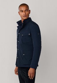 Mand iført en marineblå jakke med klaplommer og sølvknapper, stående mod en ensfarvet lys grå baggrund, og ser til venstre.