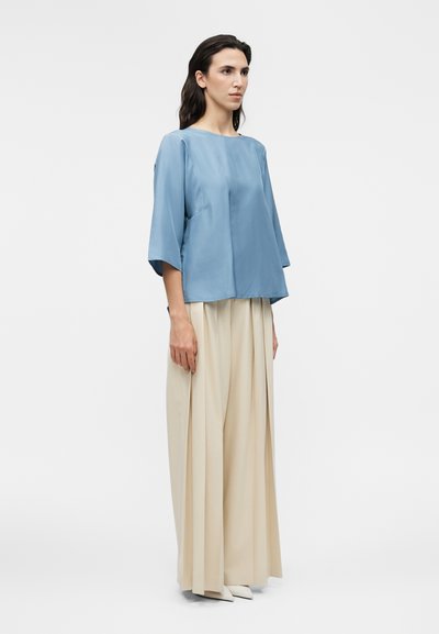 Γυναίκα που στέκεται, φοράει ένα άνετο ανοιχτό μπλε τοπ με μανίκια μέχρι τον αγκώνα και φαρδιά μπεζ πλισέ παντελόνια με λευκά μυτερά παπούτσια.