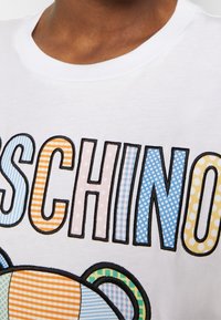 Close-up van een wit shirt met het woord "MOSCHINO" in meerkleurige, met patronen bedrukte letters en een deel van een berenafbeelding onder de tekst.