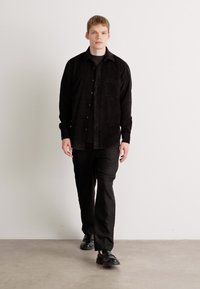 Young man walking forward wearing a black button-up shirt, black pants, and black shoes against a plain white wall.