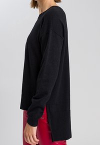 Pull noir à manches longues avec une coupe décontractée, ourlet côtelé et fentes latérales, exposé à côté de pantalons rouges brillants. Texture douce, design décontracté.