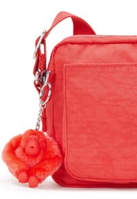 Koralfarvet crossbody taske lavet af struktureret stof, med en sølvfarvet hardware-nøglekæde og et fluffy rødt abe-charme fastgjort.
