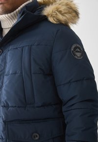 Chaqueta de plumas azul marino con capucha con ribete de pelo, que presenta un parche redondo en la manga, textura acolchada y un bolsillo frontal con cierre de botón.