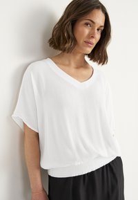 Witte, gestructureerde blouse met een V-hals en losse mouwen, met een gathered tailleband, gecombineerd met zwarte onderkleding. Zacht materiaal, casual ontwerp.