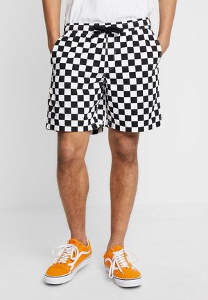 Homme portant un short à carreaux noir et blanc avec un cordon noir et des baskets orange vif avec des lacets blancs, debout les mains dans les poches.