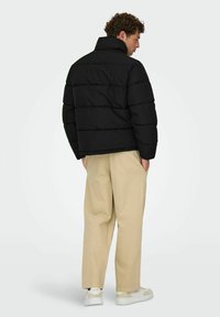Hombre con cabello rizado, vistiendo una chaqueta acolchada negra, pantalones holgados beige y zapatillas blancas, de pie con las manos en los bolsillos frente a un fondo blanco.