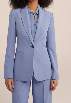 Vrouw die een lichtblauw op maat gemaakt jasje en bijpassende broek draagt, met een overhemd met kraag eronder, staand tegen een effen achtergrond.