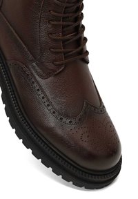 Botte en cuir marron avec tige texturée, fermeture à lacets, détails de brogue sur l'orteil et semelle en caoutchouc noir robuste.