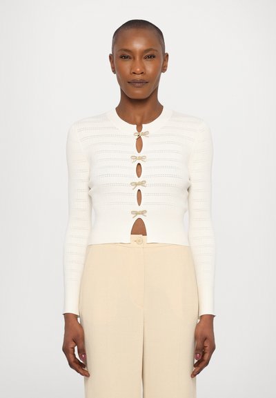 Cardigan blanc à côtes avec un col rond, doté d'accents en forme de nœud doré et de fermetures à boutons. Associé à un pantalon beige taille haute.
