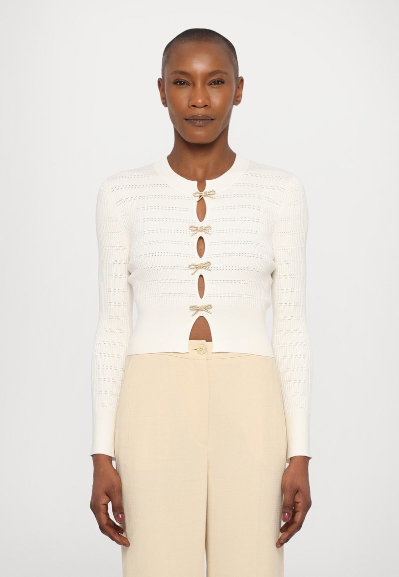 Hvit ribbestrikket cardigan med rund halslinje, som har gullfargede sløyfeakenter og knappestenginger. Kombinert med beige høytlivsbukser.