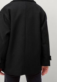 Abrigo de lana negro con un ajuste estructurado, que presenta un cuello simple, mangas largas y detalles de botones en los puños. Tela texturizada visible.