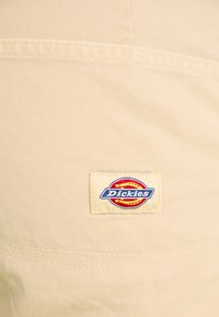 Beige cotton fabric with a smooth texture. Features a colorful Dickies logo label stitched onto the side. Simple, durable design.
