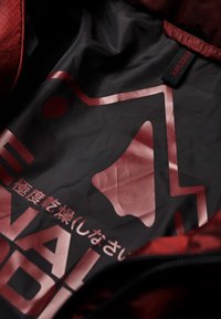 Vnitřek červené bundy s černou podšívkou s lesklým grafickým designem, který obsahuje ikonu zvonku a text tučnými červenými znaky.