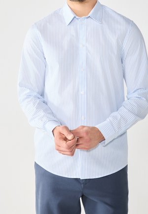 Mann trägt hellblau gestreiftes Hemd mit Knöpfen und dunkelblaue Hose, der mit beiden Händen den Manschettenknopf befestigt.