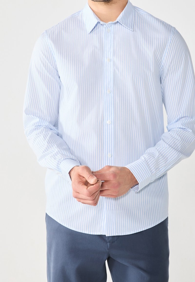 Homme portant une chemise à rayures bleu clair avec boutons et un pantalon bleu foncé, en train d'attacher le bouton de la manche avec les deux mains.