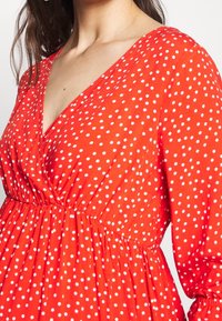 Vestido rojo con lunares blancos, que presenta un escote en V profundo, mangas largas y una cintura fruncida. La tela parece ligera y suave.