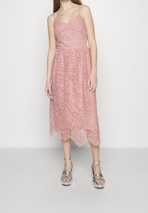 Vrouw die een mouwloze roze kanten midi-jurk draagt met dunne bandjes en zilveren plateauhoge hakken sandalen, staand tegen een effen achtergrond.