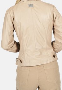 Béžová kožená bunda s přiléhavým střihem, která má štítek na krku, boční přezky a viditelné švy na zadní straně.