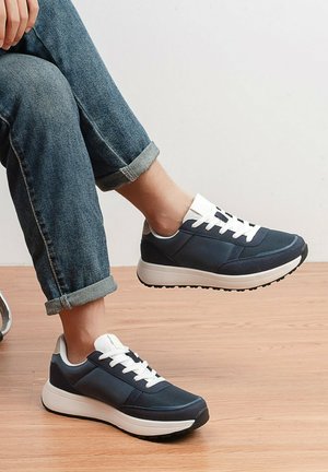 Personne portant un jean bleu retroussé et des baskets bleu marine et blanches, assise avec un pied levé au-dessus d'un sol en bois.