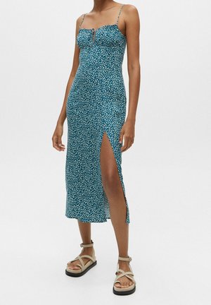 Kvinde iført teal slip dress med hvidt blomsterprint og højt slids, kombineret med beige sandaler med stropper og platform.