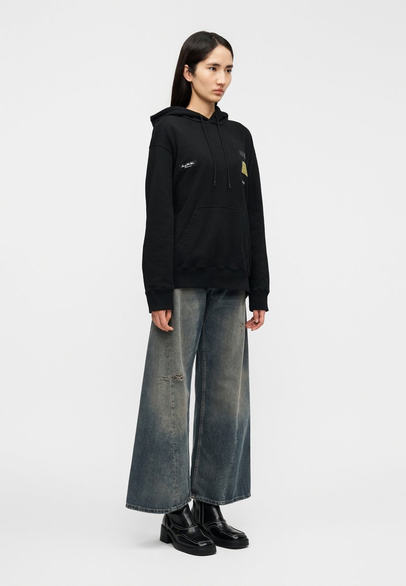 Black hooded sweatshirt with front pocket, graphic details. Paired with wide-leg faded blue jeans featuring rips. Black platform shoes.