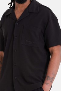 Zwart kortärmige shirt van een gestructureerde stof, met een knoopsluiting en een borstzak met subtiele branding. De knopen zijn donker.