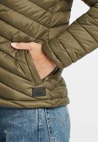 Olivenfarget polstret jakke med chevron-quilting, med en sidelomme og en svart etikett. Teksturen er glatt og skinnende.