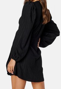 Vestido negro con mangas largas abullonadas, textura acanalada, cintura ajustada y corte relajado. Presenta un corte corto y un brillo sutil.